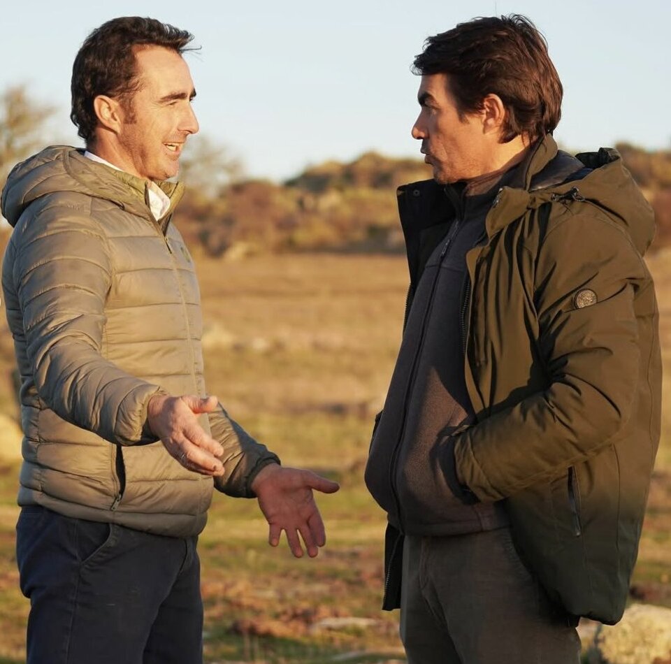 Dos hombres conversando en un campo, uno con chaqueta gris y otro con chaqueta verde.