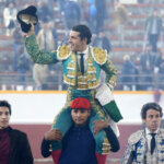Torero levantando el sombrero mientras es llevado en hombros por un hombre.