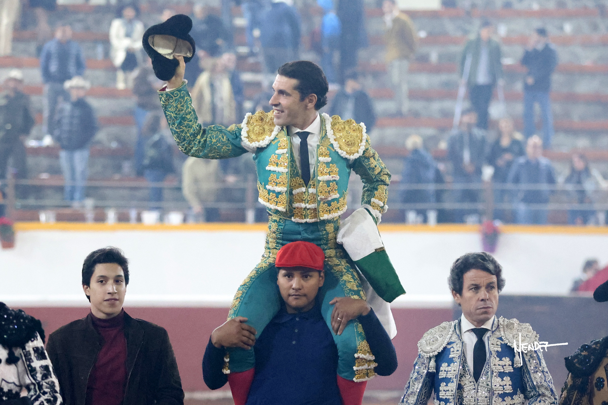 Torero levantando el sombrero mientras es llevado en hombros por un hombre.